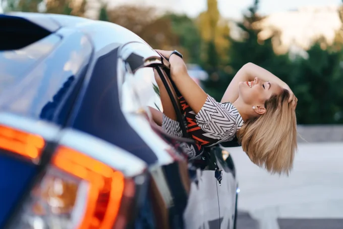 Cheerful Caucasian woman drives through the picturesque sunny city and waves her arms while stretching out of the car window on a beautiful day